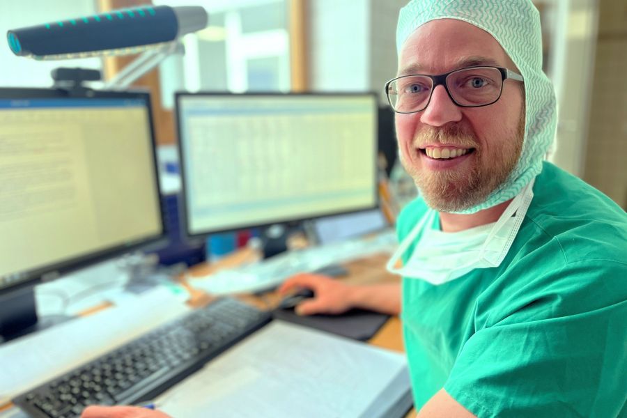 Patrick Dorsten sitzt in OP-Kleidung am Schreibtisch und schaut in die Kamera. 
